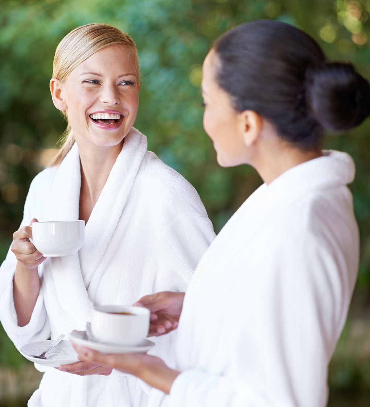 Two women in white robes enjoying coffee and chatting outdoors.